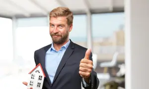 Ein Mann im Anzug hält ein Hausmodell und zeigt Daumen hoch. Er ist ein Investor, der in Immobilien investiert.