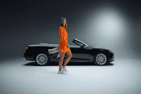 Frau in orangefarbenem Kleid neben schwarzem Cabrio. Ein toller Sixt Aktie Rabatt für Ihre nächste Fahrt!