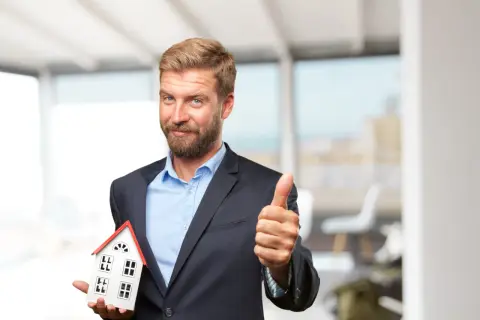 Ein Mann im Anzug hält ein Hausmodell und zeigt Daumen hoch. Er ist ein Investor, der in Immobilien investiert.